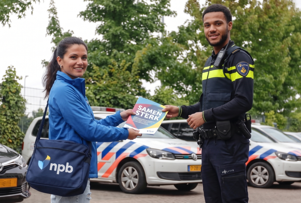 Woman in blue jacket hands a brochure to a uniformed officer outdoors, smiling with police cars in background.