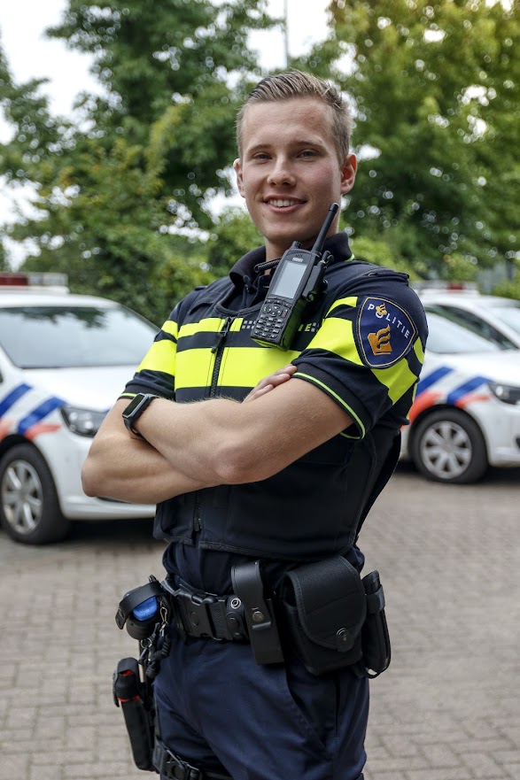 Police officer in uniform with radio and gear, standing with arms crossed in front of squad cars outdoors.