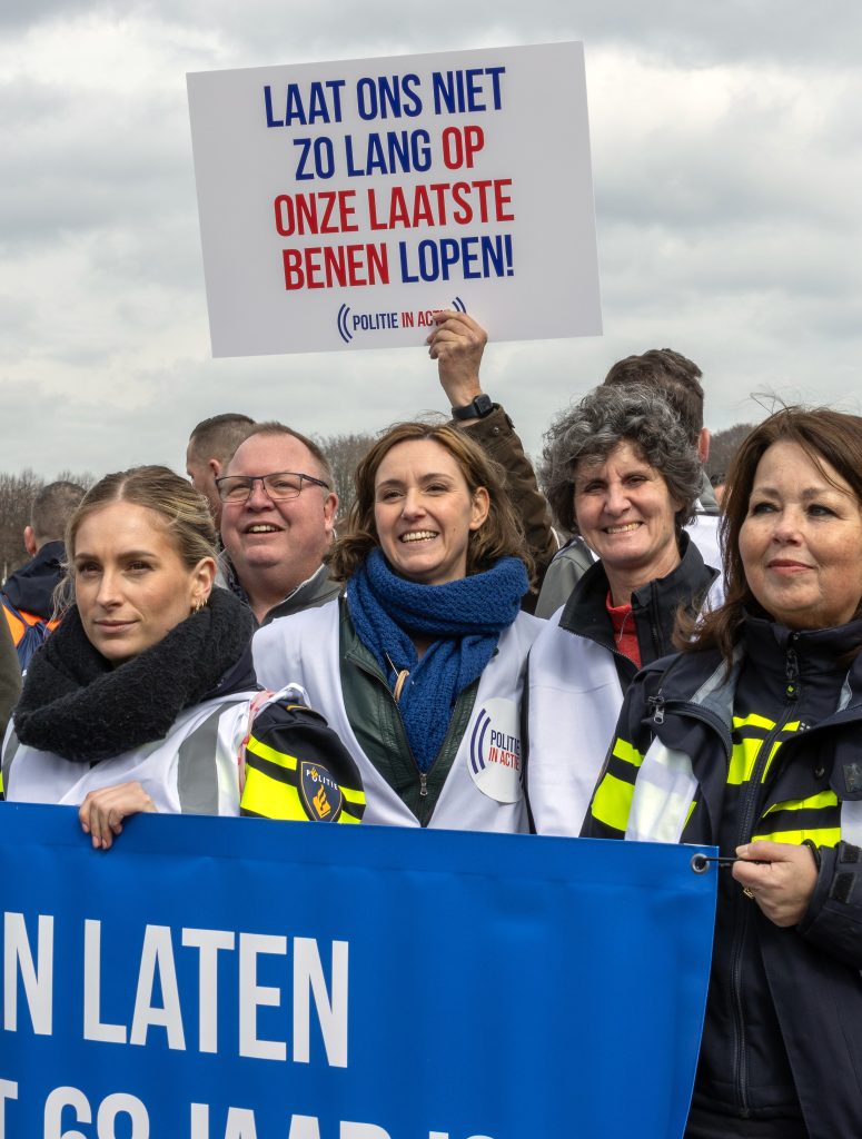 Group of protesters holding a large blue banner with white letters, smiling at the camera; Dutch slogan on sign reads 'Laat ons niet zo lang op onze laatste benen lopen' (We won’t walk for long on our last legs).