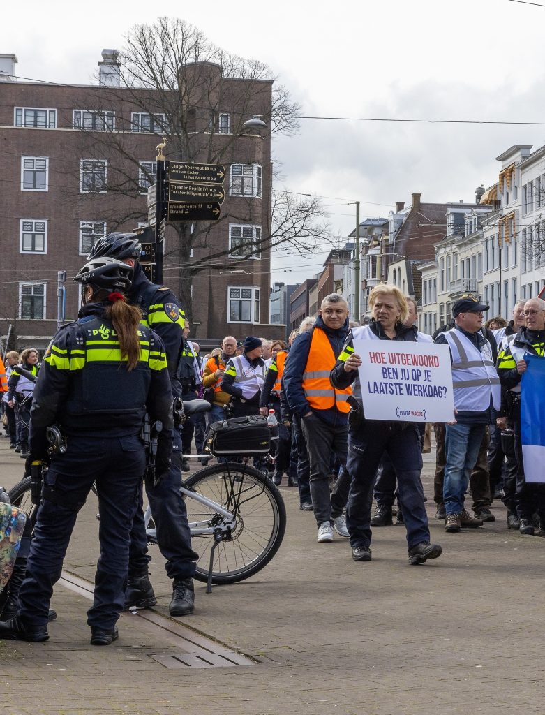 Crowd of protesters wearing high-visibility vests standing on a city street, with police officers and a bicycle in the foreground and buildings in the background.