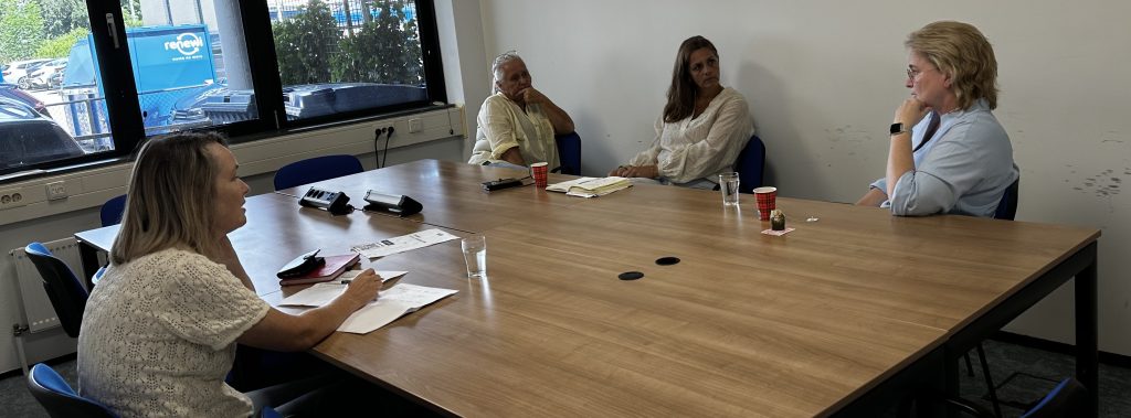 Four women sit around a wooden conference table in a meeting room, discussing with papers and cups on the table.