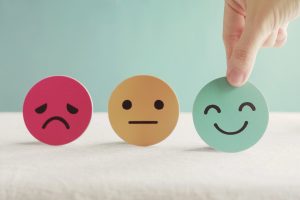 Three colorful emoji faces in a row (pink sad, beige neutral, green happy) with a hand touching the green smiley on the right.