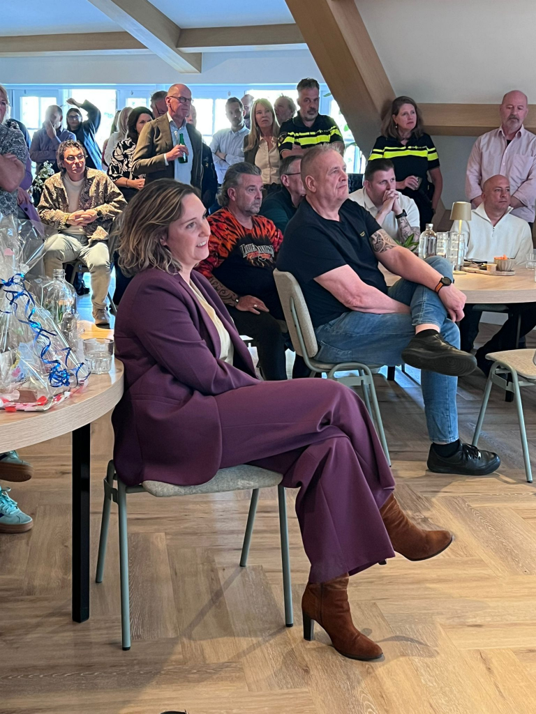 Group of people at a casual indoor gathering; a woman in a purple suit sits in the foreground while others stand and chat in the background (gift baskets on a table to the left).
