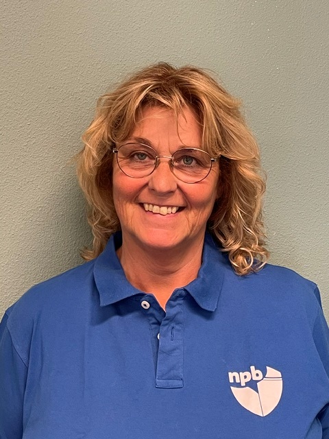 Smiling woman with blonde hair and glasses in a blue polo shirt with an NPB logo on the left chest.