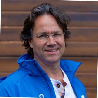 Man with brown hair and glasses wearing a bright blue jacket, standing in front of wooden steps and smiling slightly.