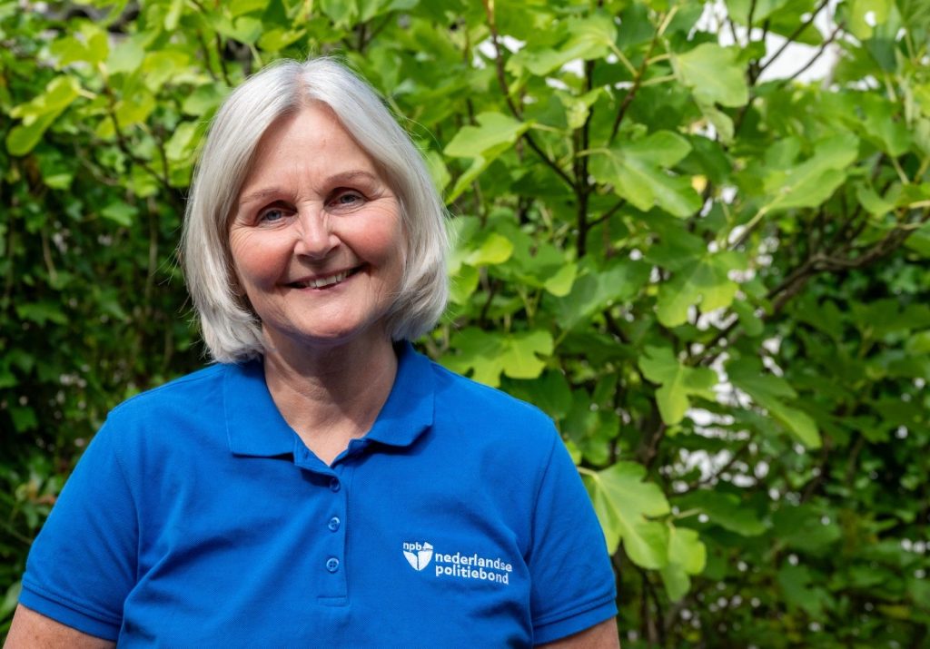 Smiling older woman with short gray hair wearing a blue polo shirt, standing among green bushes in sunlight.