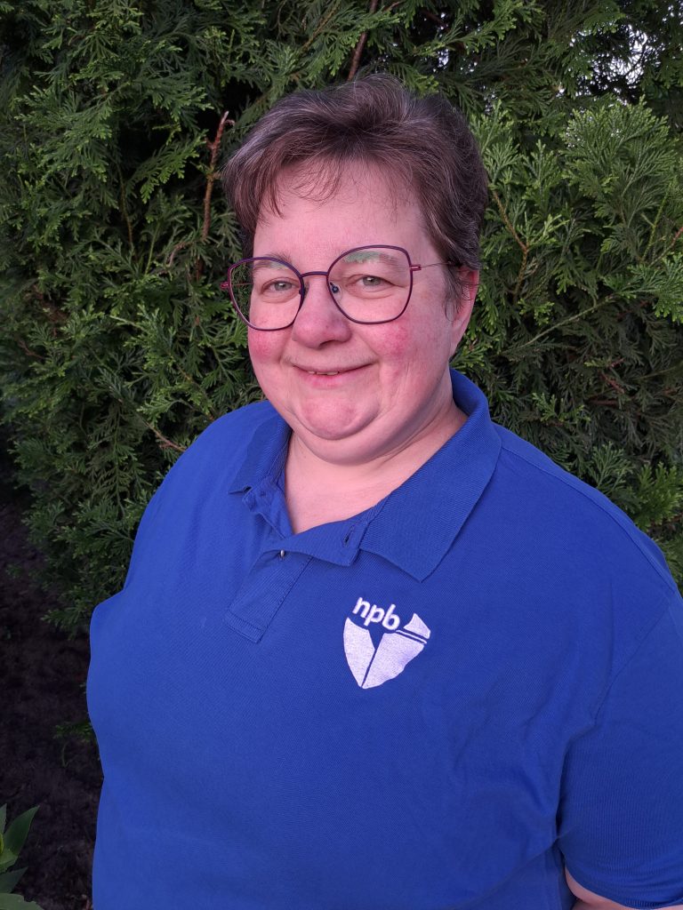 Person with short hair and glasses wearing a blue polo shirt with an HP logo, standing in front of a green evergreen hedge outdoors, smiling at the camera.