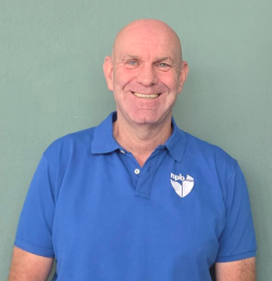 Smiling bald man wearing a blue polo shirt with a white logo on the chest, standing in front of a green wall.