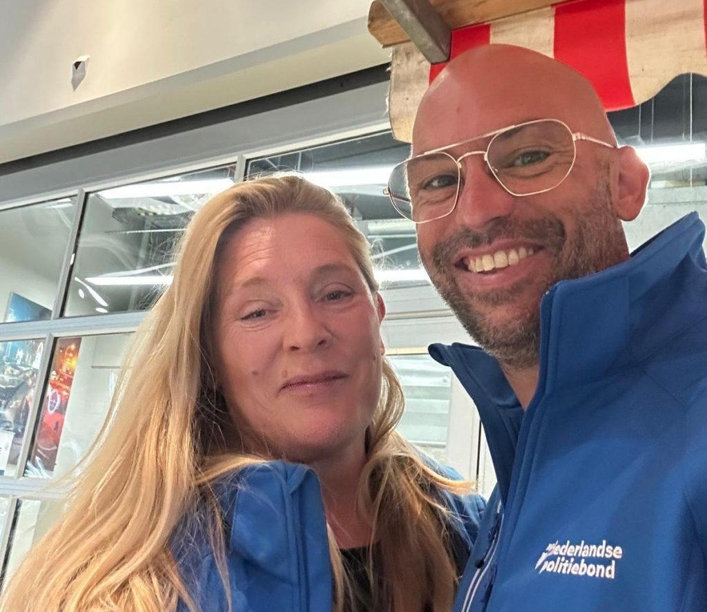 Two people in blue jackets take a cheerful selfie indoors, smiling at the camera.