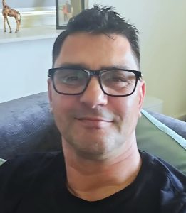 Close-up portrait of a man with short dark hair and black rectangular glasses, smiling gently while seated on a gray couch wear­ing a black shirt.