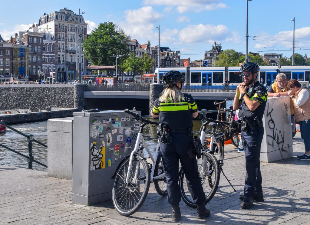 Politie aan het werk in de zomer FOCUS2 - Nederlandse Politiebond Two cyclists in high-visibility uniforms stand with their bikes by a waterfront railing, with a blue tram and historic buildings in the background.