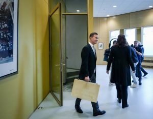 Man in a dark suit holding a beige briefcase or folder, standing in a hallway with other people in the background.