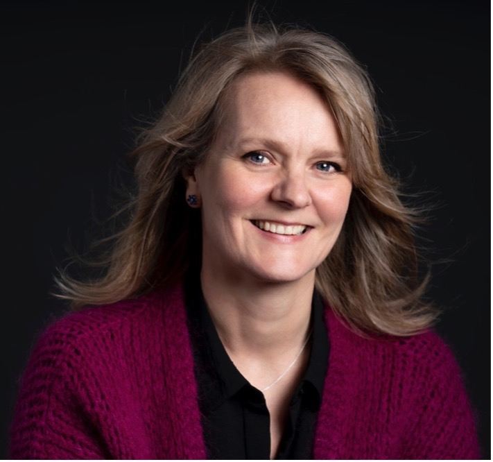 Smiling woman with light brown hair, wearing a magenta cardigan and black blouse against a dark background.