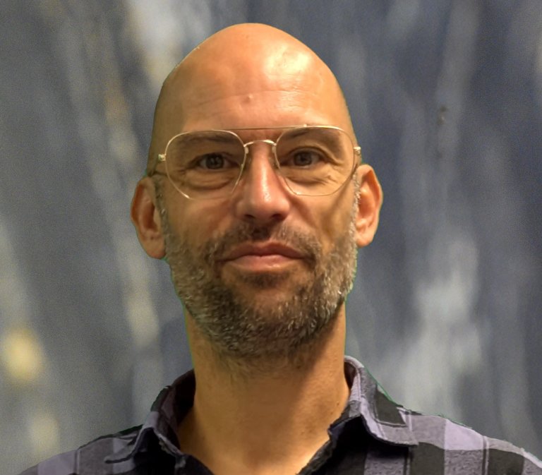 Portrait of a bald man with round glasses and light stubble, wearing a blue-gray plaid shirt against a blurred background.