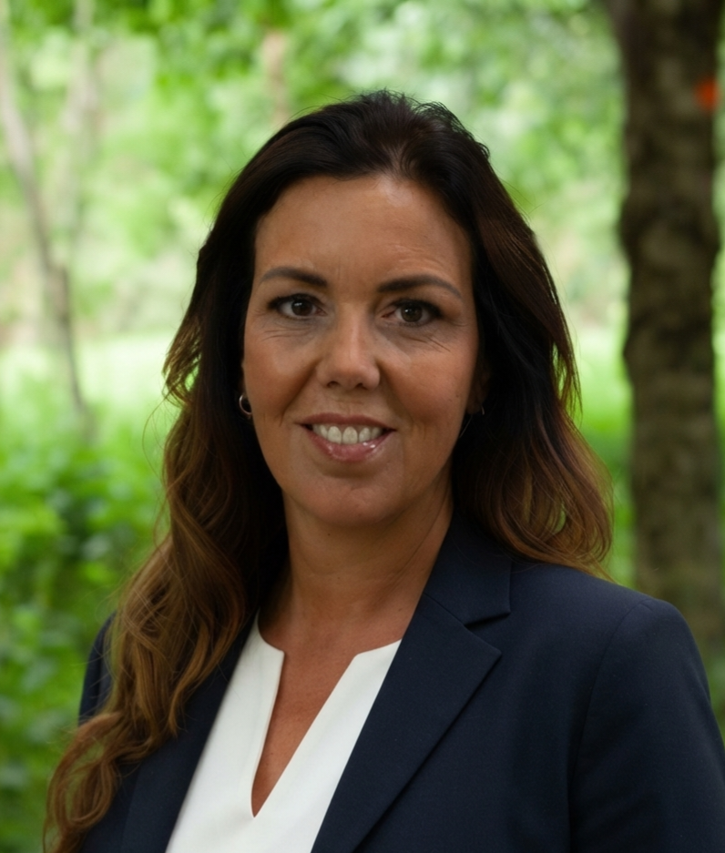Professional woman smiling outdoors in a navy blazer with a white blouse, greenery in the background.