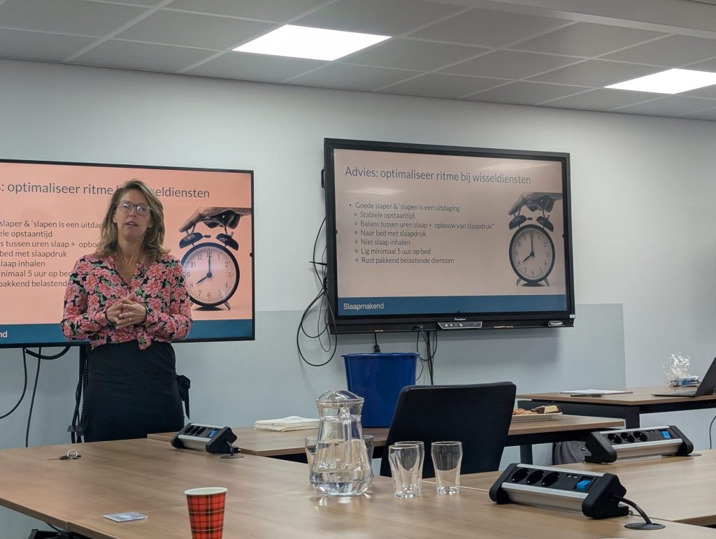 Female presenter in a floral top standing beside orange posters in a meeting room.