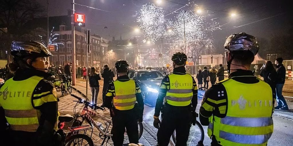 politie-jaarwisseling-v4 - Nederlandse Politiebond Nighttime street scene with Dutch police officers in neon jackets and bicycles, watching fireworks and a crow d of bystanders.