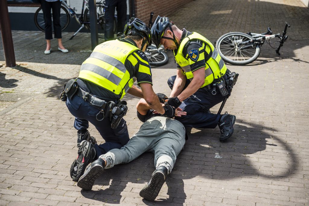 Two police officers in high-visibility vests help a person who is lying on the pavement, surrounded by bicycles.