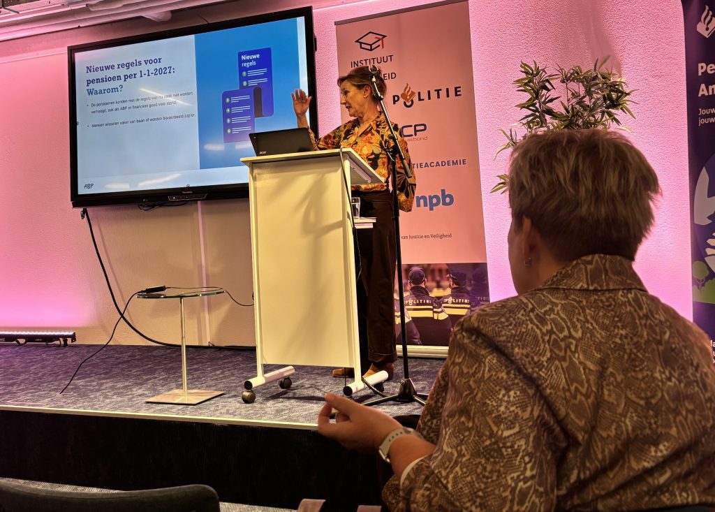 Foto Politiepensioen Baarn A OBLONG FOCUS - Nederlandse Politiebond Presenter at a podium giving a talk; a large slide about pension rules in Dutch is on screen, audience visible in foreground.