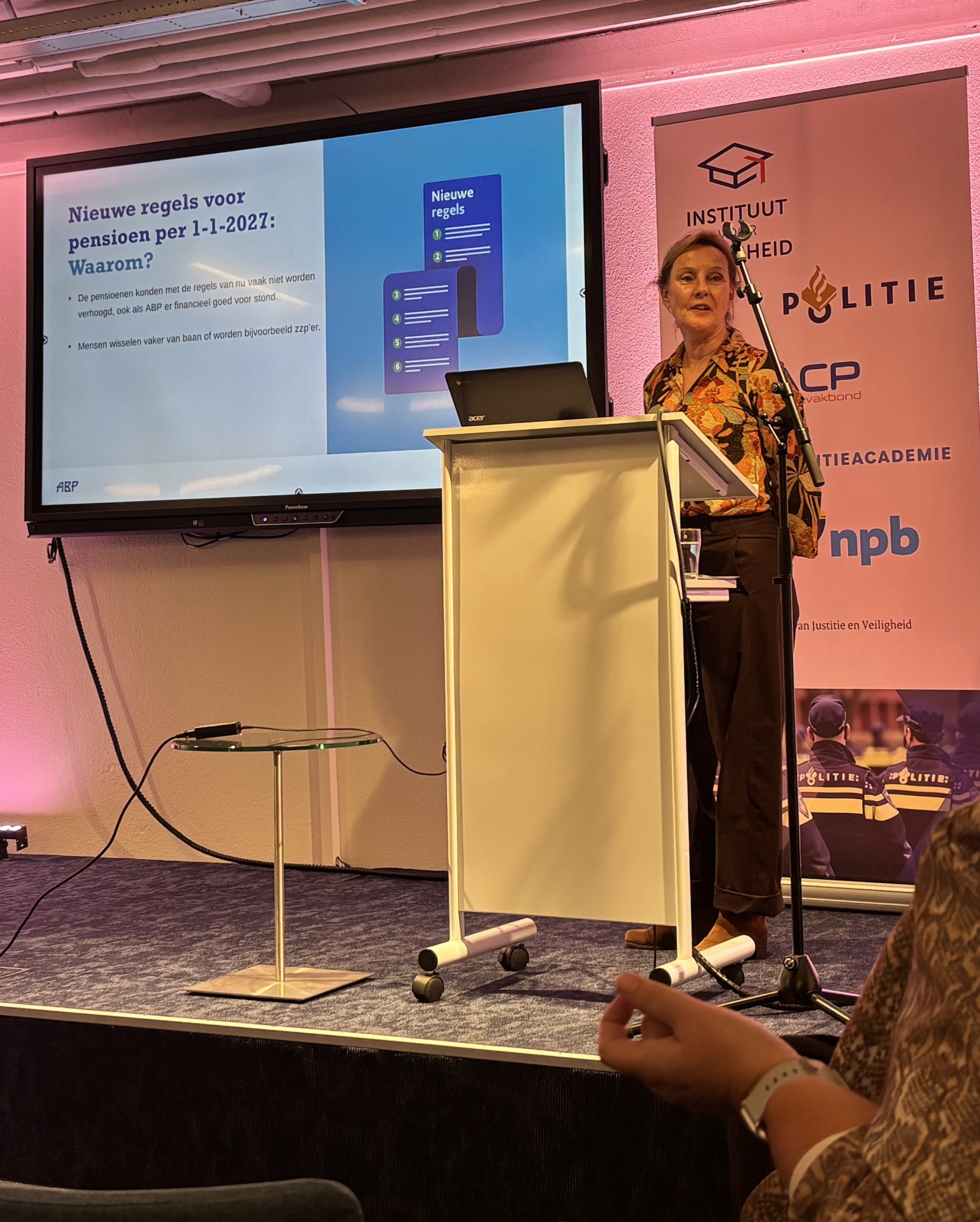 Presenter at a lectern giving a talk; slide on pension rule changes in Dutch visible on a large screen, with a police banner in the background.
