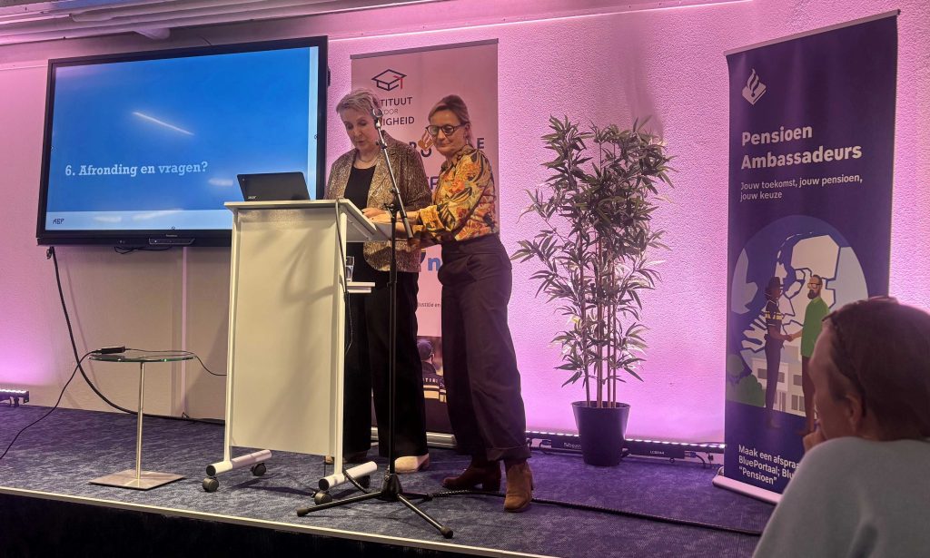 Two women speak on a stage at a podium with a laptop; a blue slide reads '6. Afronding en vragen?'