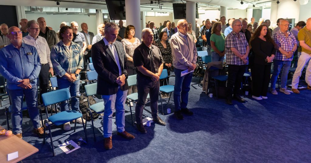 Group of adults standing in a conference room audience, facing forward, with blue chairs and a blue carpet visible.