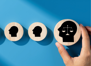 Series of wooden discs with black head silhouettes, the last held by a hand showing a head with scales of justice inside, symbolizing legal psychology or fairness in decision-making.