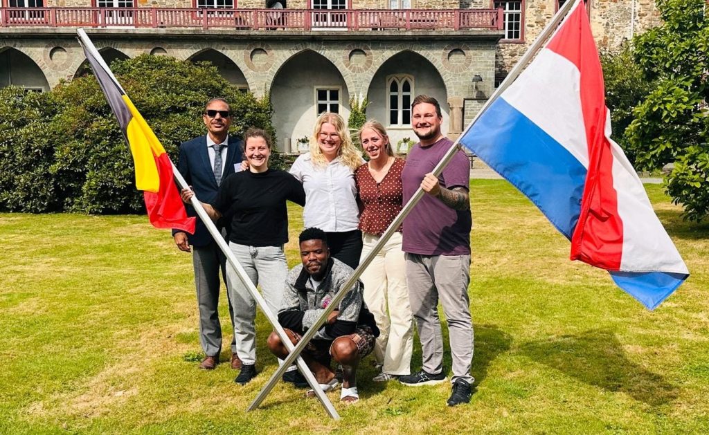 Six people pose on a lawn holding two large flags (Belgian and Dutch) in front of a historic stone building with arches and greenery.