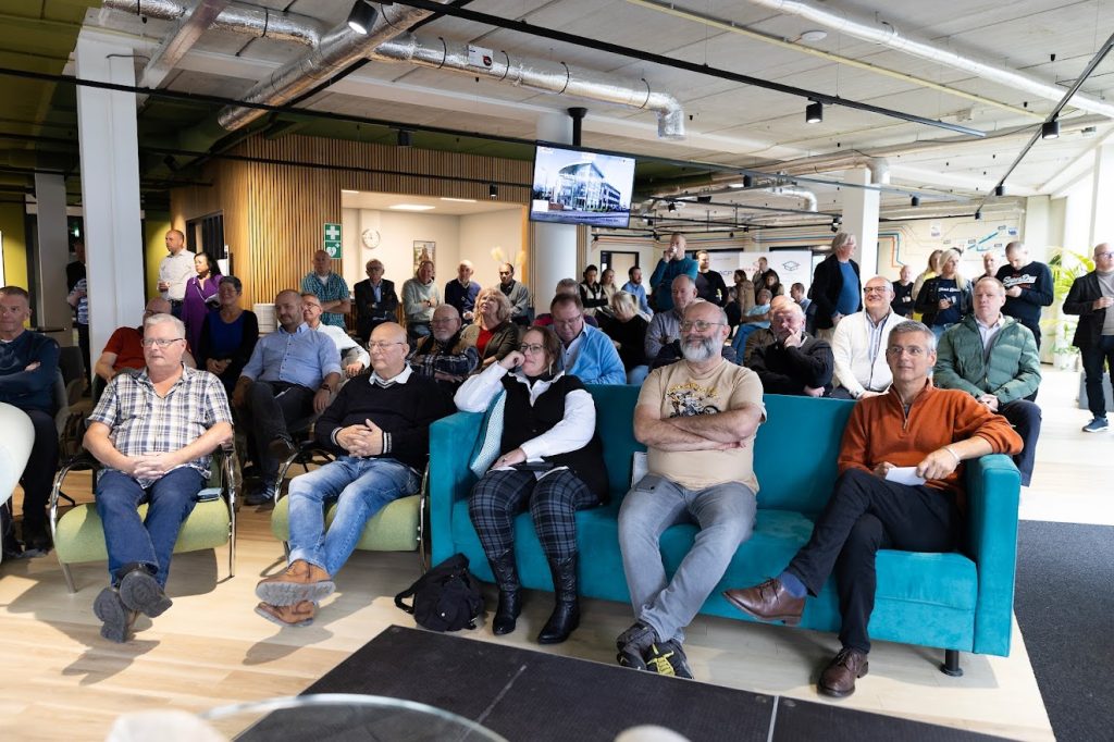 Audience seated in a bright open room watching a presentation, some on teal couch and others on chairs.