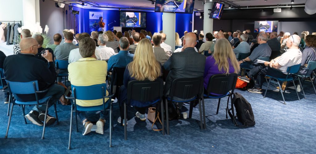Large audience seated in a conference room facing a stage with a presenter and multiple screens glowing blue-based lighting nearby