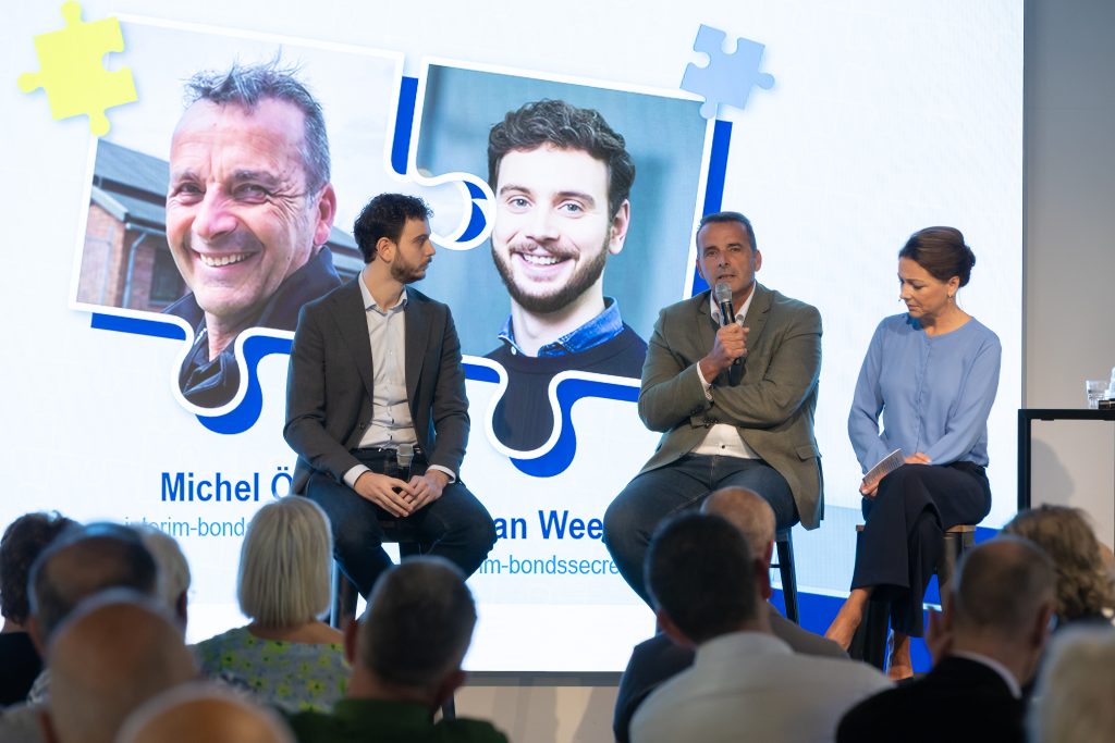 interim-db-1 - Nederlandse Politiebond Panel discussion on stage with three panelists (two men in suits and a woman in blue) in front of a large screen showing portrait photos and puzzle-piece graphics behind them.