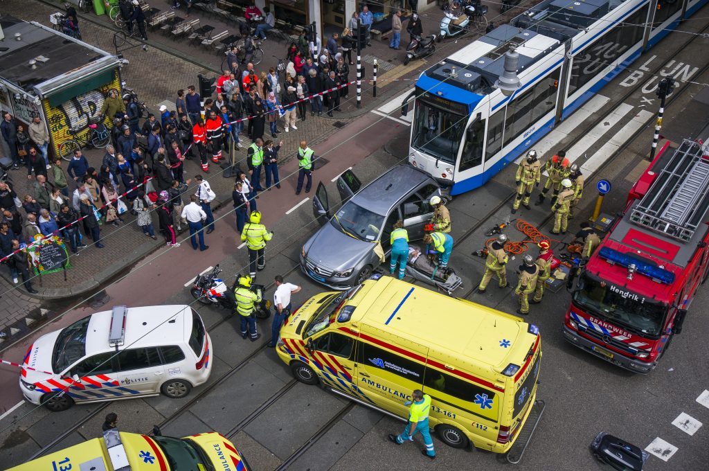 Crowd gathers on a city street as a white and blue tram is blocked by emergency crews nearby, with firefighters and police present on scene.”,